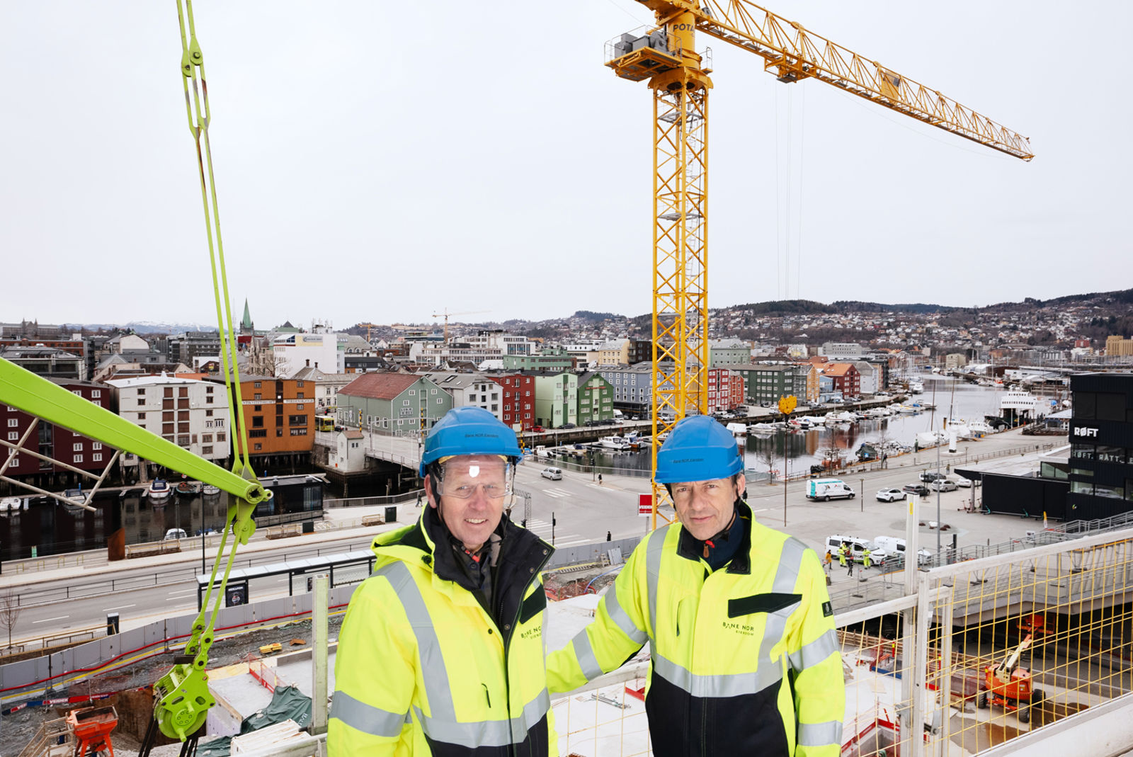to menn i arbeidstøy smiler. bak dem er det utsikt til byggeplass med heisekran.